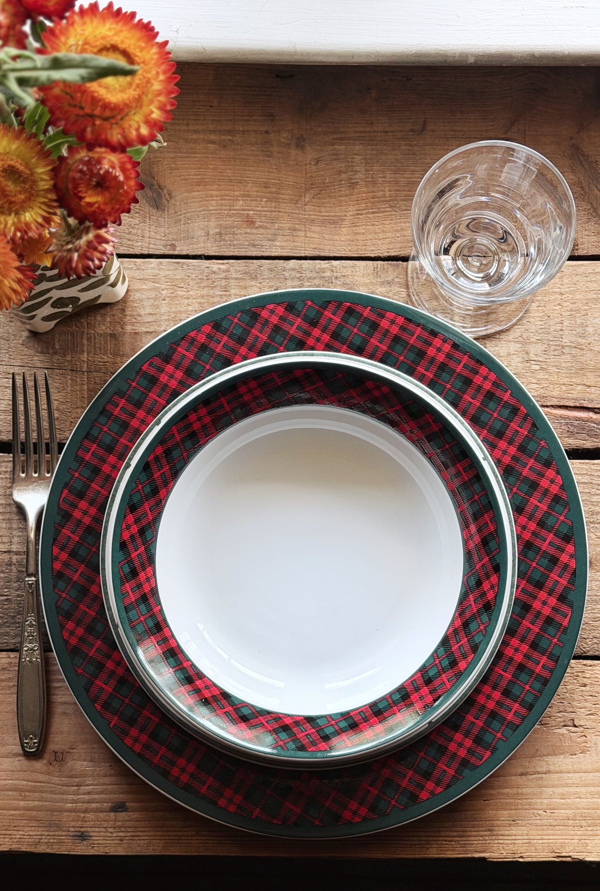 A place setting on a rustic wooden table featuring a stack of plates with a red and green plaid pattern and a white center plate, accompanied by a fork, a clear drinking glass, and dried orange flowers.