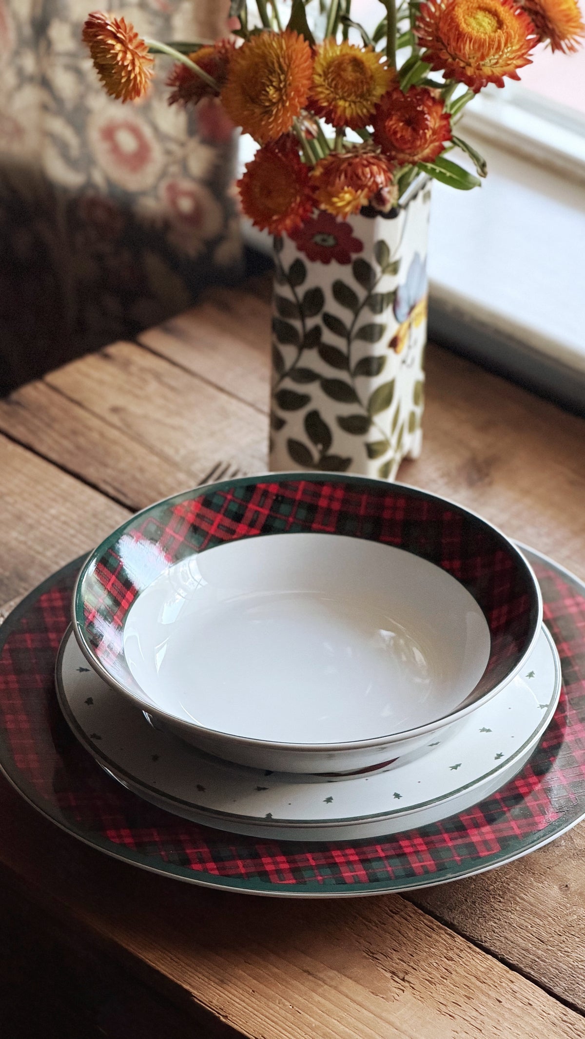 A stack of white ceramic plates with red and green plaid and speckled patterns, styled on a rustic wooden table next to a floral-patterned vase of orange flowers.