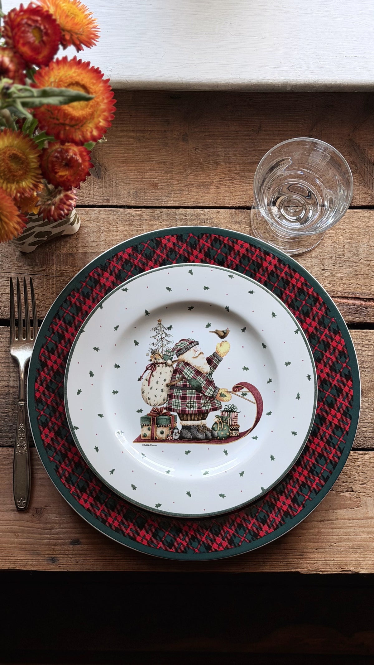 A festive holiday table setting featuring a white ceramic plate with a Christmas Santa illustration in the center, layered over a red and black plaid charger plate with a dark green rim. The setting is on a rustic wooden table with a fork, a glass, and a vase of orange flowers.