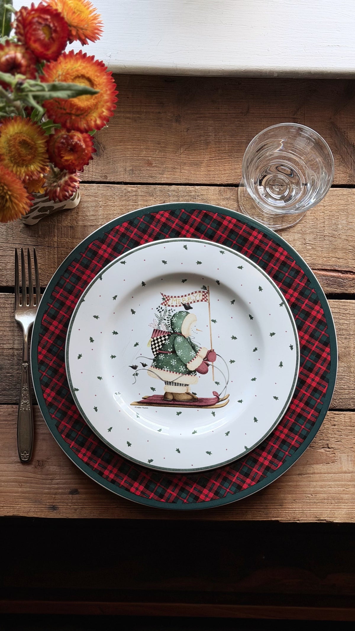 A festive holiday table setting featuring a white ceramic dinner plate with a Christmas snowman illustration, layered over a red and black plaid charger plate with a dark green rim, on a rustic wooden table.