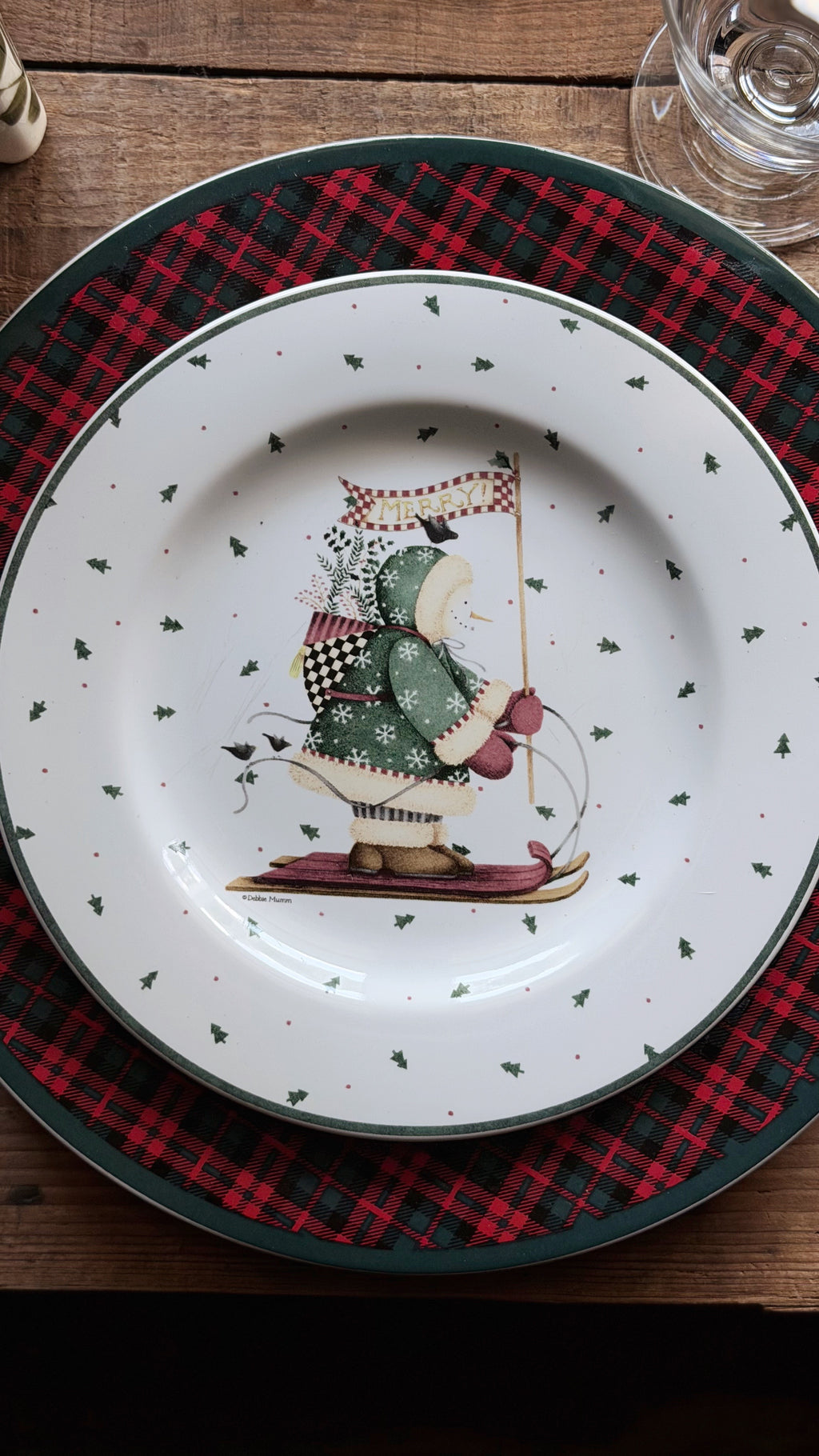 A Debbie Mumm Sakura Sledding Characters Christmas Salad Plate featuring a winter scene illustration of a person in a green coat and red hat on skates, surrounded by small green holly motifs and a dark green rim. The plate is layered on top of a red and black plaid placemat on a wooden table.