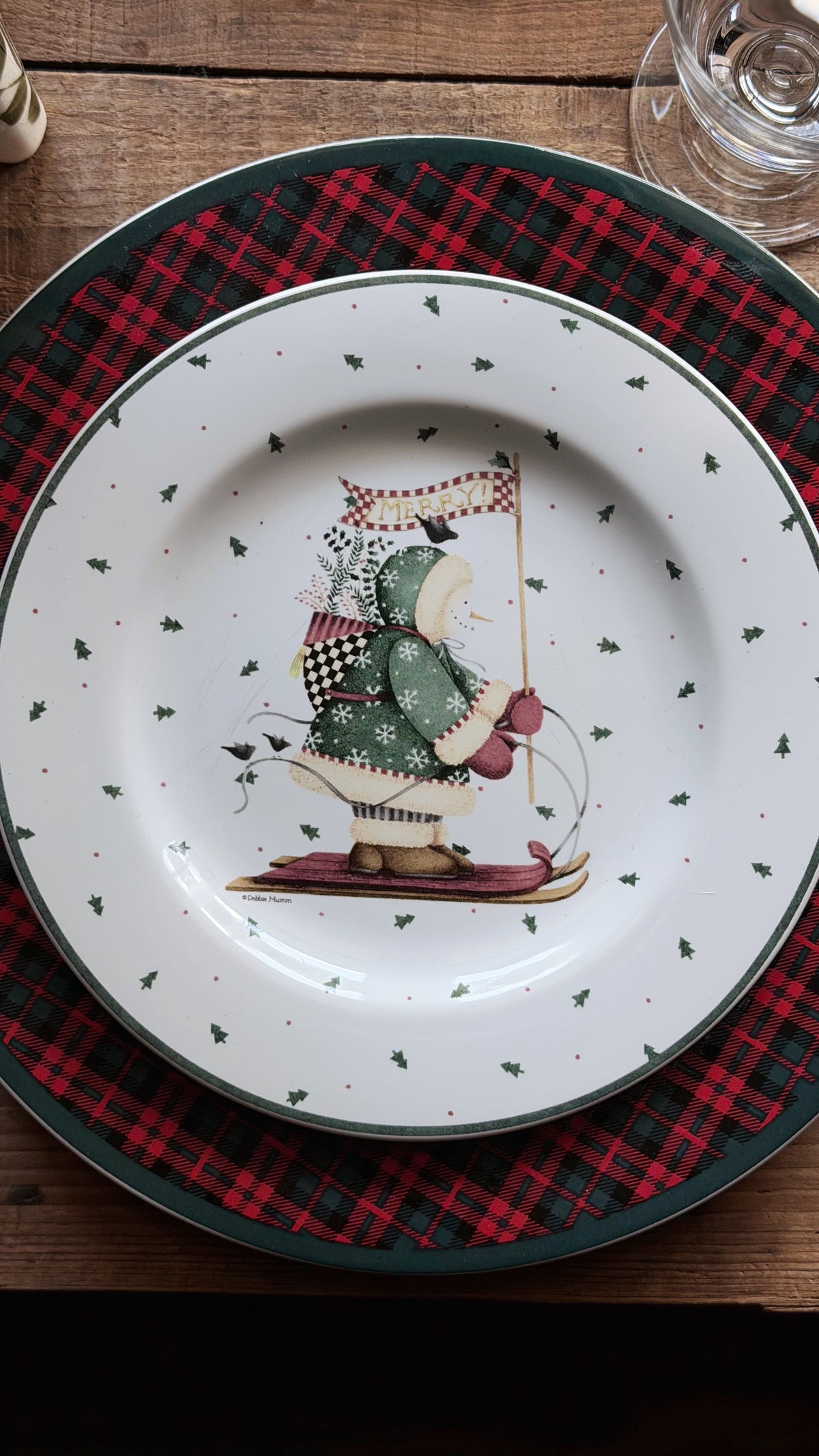 A Debbie Mumm Sakura Sledding Characters Christmas Salad Plate featuring a winter scene illustration of a person in a green coat and red hat on skates, surrounded by small green holly motifs and a dark green rim. The plate is layered on top of a red and black plaid placemat on a wooden table.