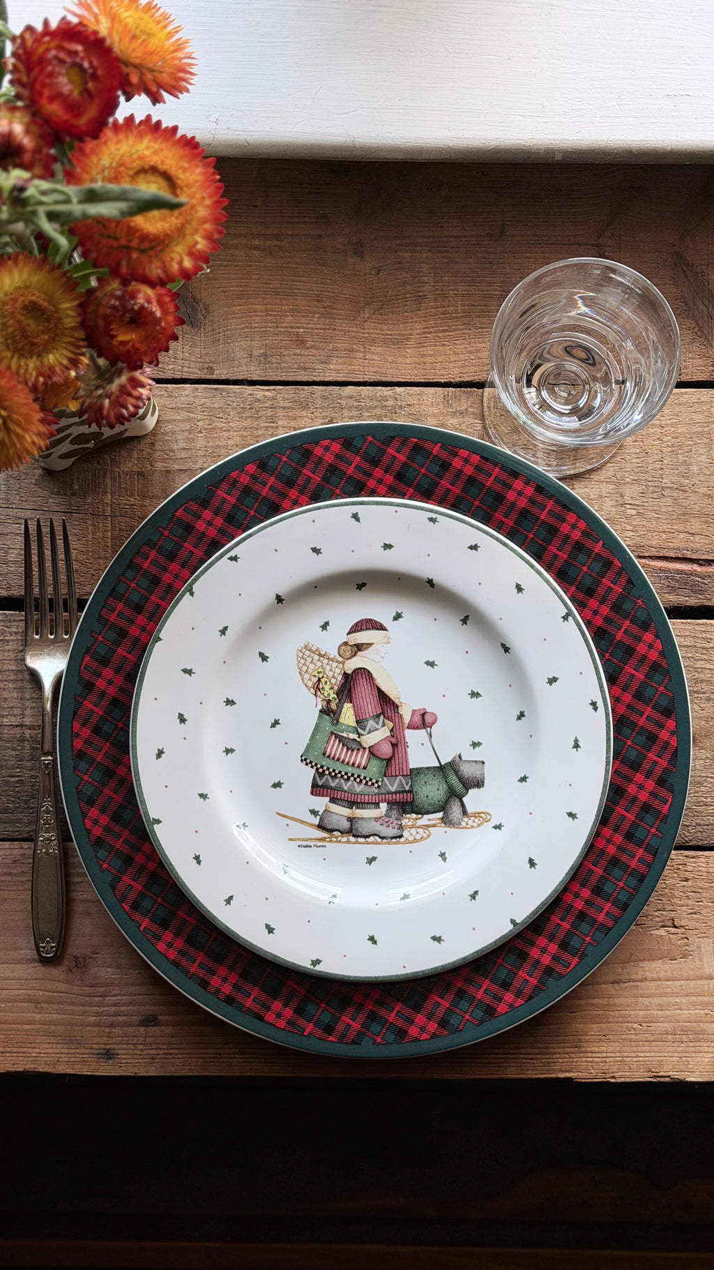 A place setting on a rustic wooden table featuring a white dinner plate with a whimsical winter character illustration in the center, layered over a red and black plaid charger plate. A silver fork, a clear drinking glass, and a small vase of orange flowers are arranged around it.