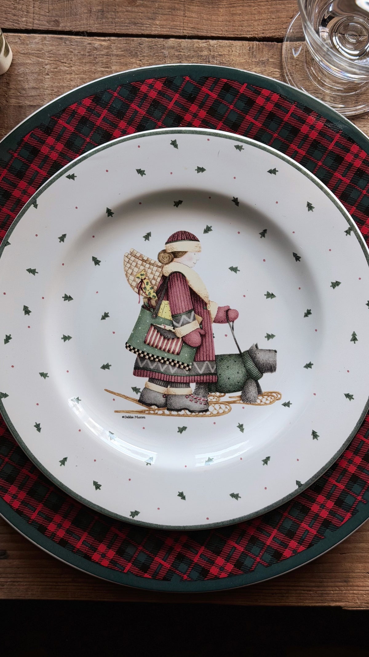 A Debbie Mumm Sakura Sledding Characters Christmas Salad Plate with a festive illustration of a woman in a winter coat and hat carrying a sack, placed on a red and black plaid charger plate over a wooden table.