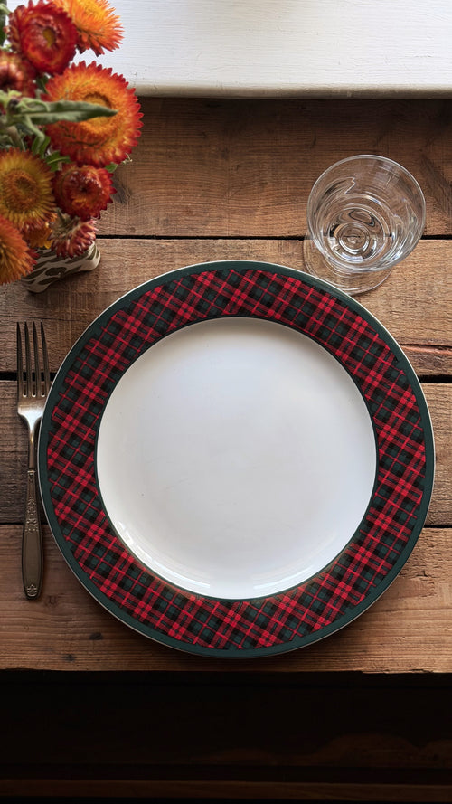 A Vintage Holiday Tartan Plaid Stoneware Dinner Plate with a red and black plaid border, set on a rustic wooden table with a fork, a clear drinking glass, and dried orange flowers.