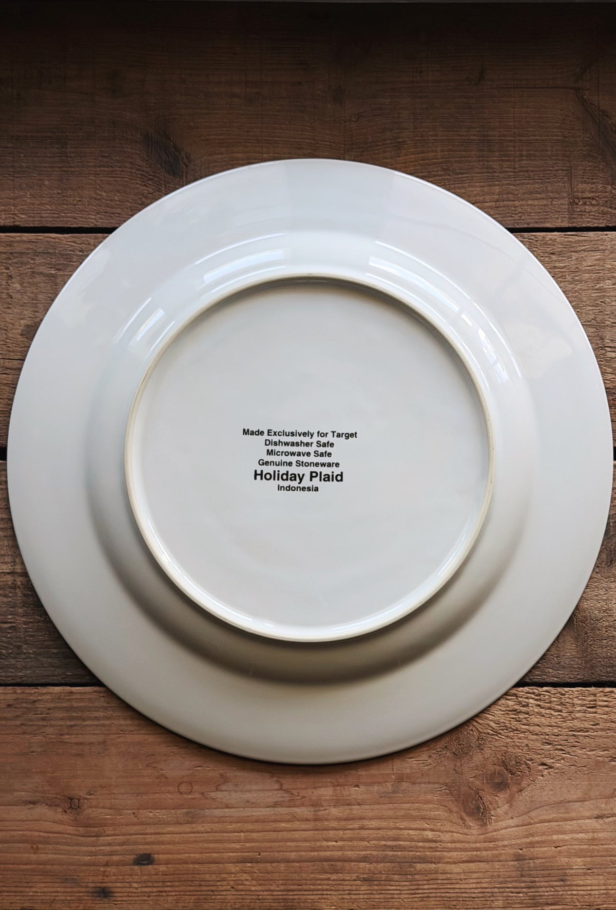 A white ceramic Christmas plate shown from the bottom side, featuring black printed text that reads 'Made Exclusively for Target Holiday Plaid Indiana.' The plate is photographed from directly above on a rustic wooden surface.