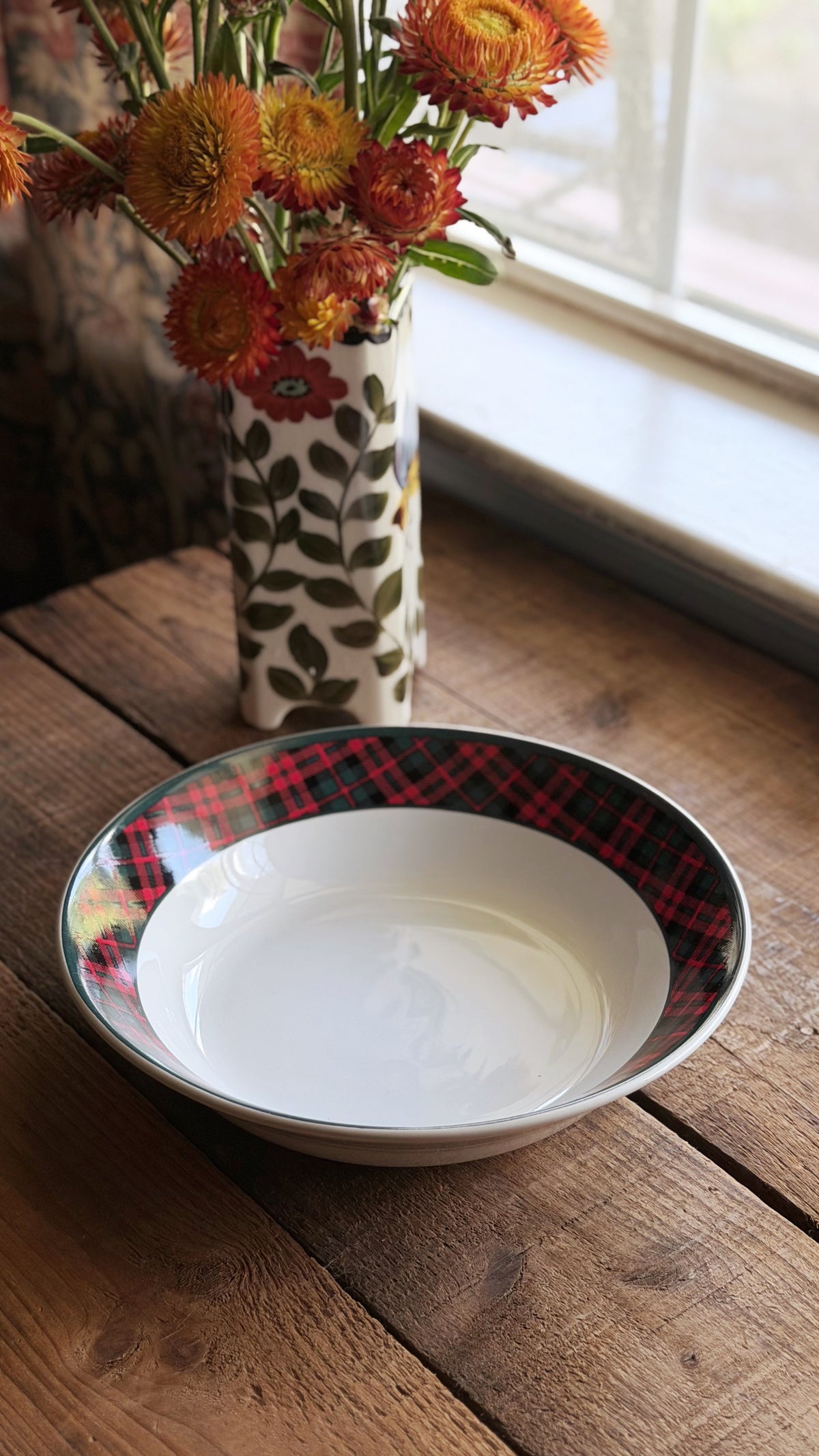 AVintage Holiday Tartan Plaid Stoneware Bowl with a white interior and a red plaid rim, set on a rustic wooden table next to a window with a floral-patterned vase of orange flowers in the background.