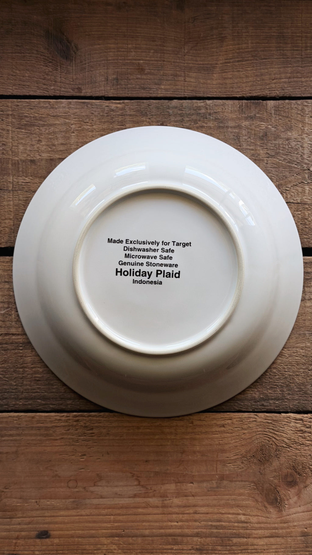A white ceramic plate shown from the bottom side, featuring black printed text that reads 'Made Exclusively for Target Dishwasher Sale Microwave Sale Genuine Stoneware Holiday Plaid Indonesia', placed on a rustic wooden surface.