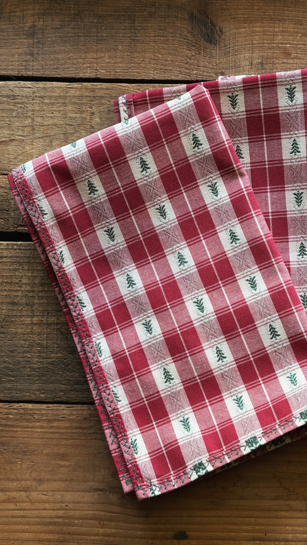 A folded red and white plaid napkin with small green pine tree motifs, displayed on a rustic wooden surface.