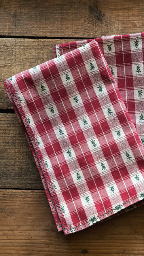 A folded red and white plaid napkin with small green pine tree motifs, displayed on a rustic wooden surface.