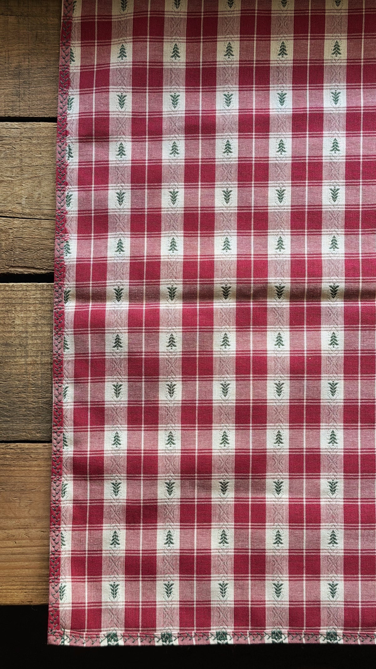 A vintage napkin with a red and white gingham plaid pattern, decorated with small green pine tree motifs, laid flat on a rustic wooden surface.