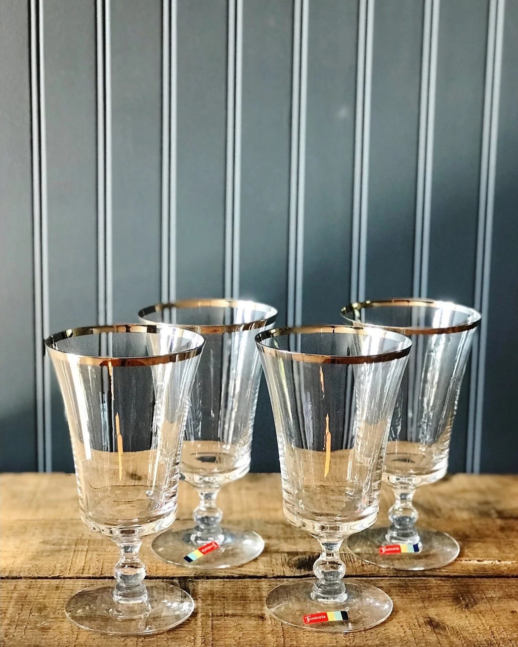 Four clear crystal or glass goblets with gold rim detailing, arranged on a rustic wooden surface against a dark gray paneled wall.