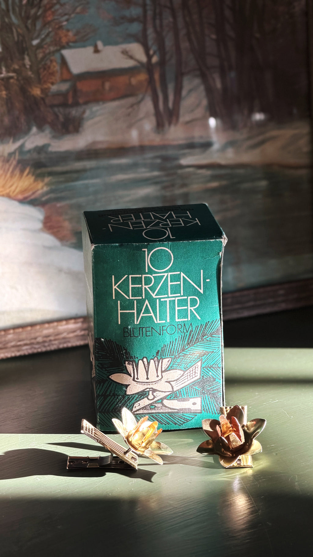 A vintage teal and green box of Kerzen-Halter candle clips sits on a dark surface with two flower-shaped candle clips placed in front. The background shows a softly blurred painted winter landscape.