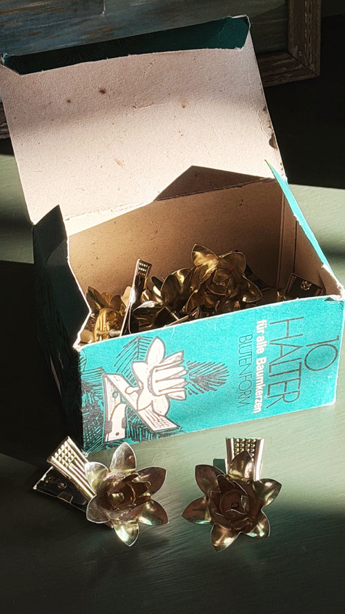 A vintage teal and white box of candle clips sits open on a dark surface, with two flower-shaped metal candle clips placed in front. The clips appear gold-toned, and the box lid is propped open behind them.