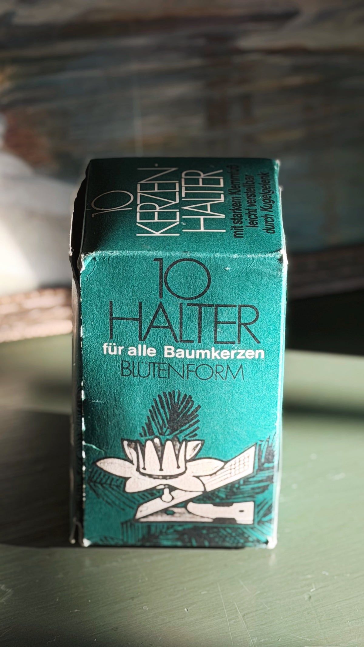 A teal-green rectangular box of flower-shaped tree candle clips, with German text '10 HALTER' and 'für alle Baumkerzen BLUTENFORM', standing on a green surface with a blurred rustic wooden background.