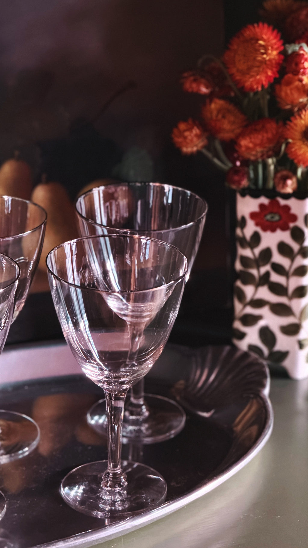 Clear crystal wine glasses arranged on a silver tray, with a dark background and a vase of orange flowers in the rear.