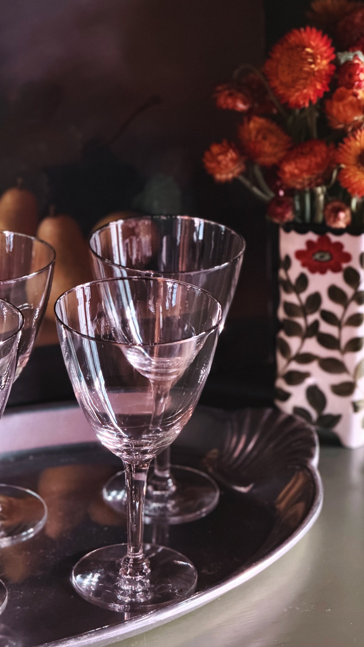 Clear crystal wine glasses arranged on a silver tray, with a dark background and a vase of orange flowers in the rear.