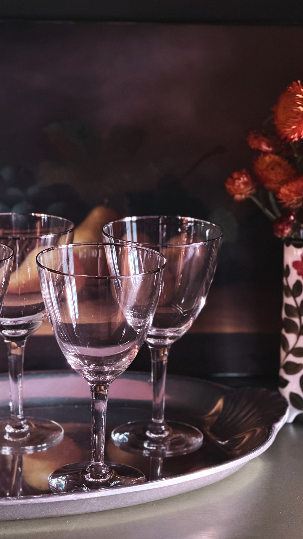 Three clear stemmed wine glasses arranged on a round silver tray, with a softly blurred dark background and orange flowers in the corner.