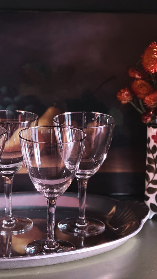 Three clear stemmed wine glasses arranged on a round silver tray, with a softly blurred dark background and orange flowers in the corner.