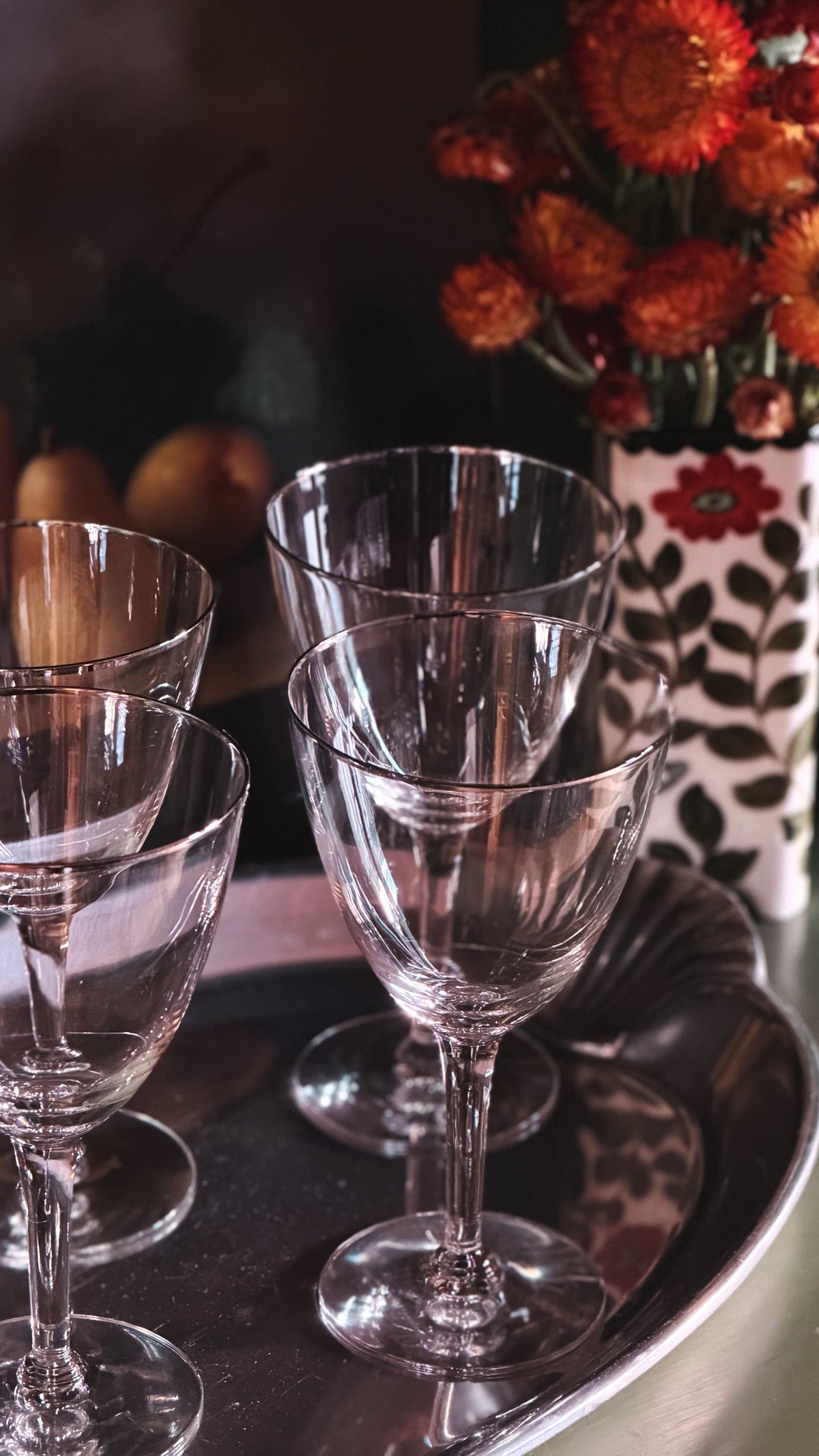 A set of clear glass wine glasses arranged on a dark metal tray, with a floral-patterned vase of orange flowers blurred in the background.