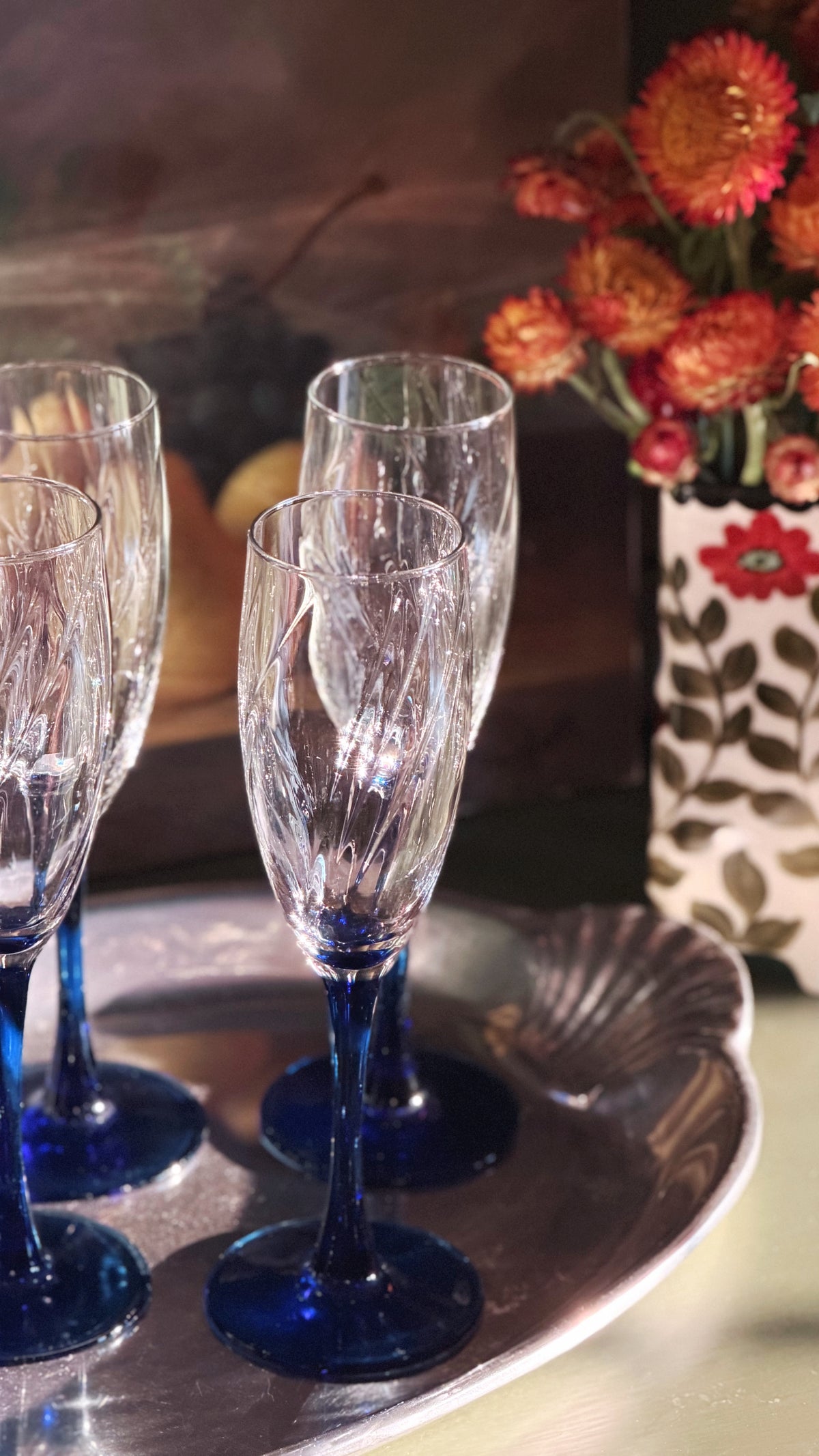 Three clear champagne flutes with etched vertical patterns and cobalt blue stems and bases, arranged on a silver tray with dried flowers in the background.