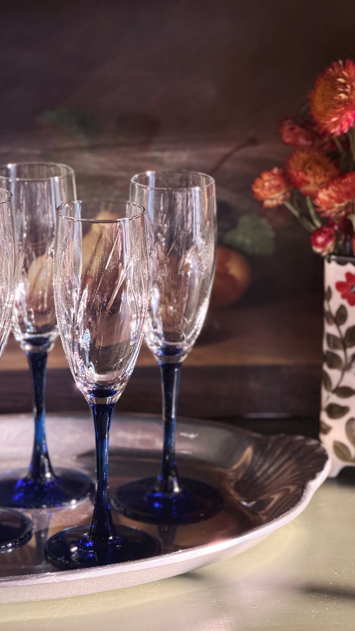 Four clear champagne flutes with dark cobalt blue stems and bases, arranged on a silver tray with a blurred floral background.