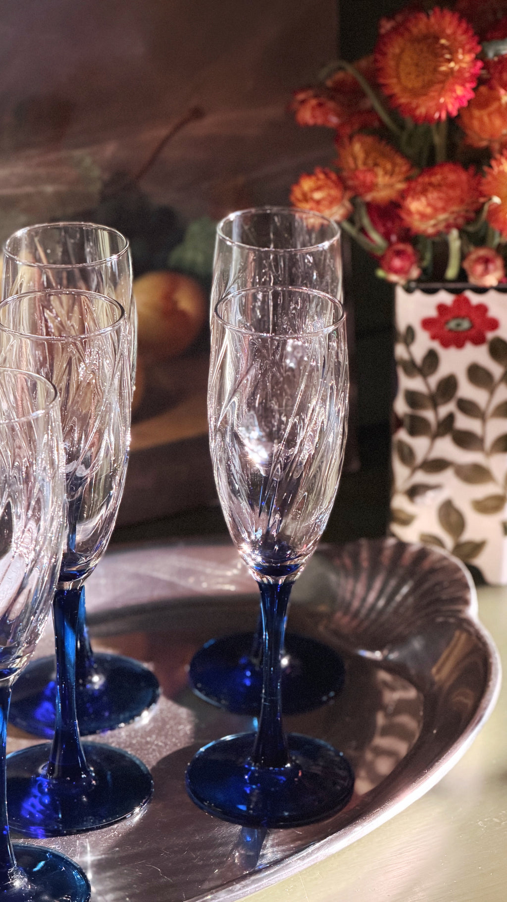 A set of clear champagne flutes with deep cobalt blue stems and bases, arranged on a silver tray with dried orange flowers in the background.