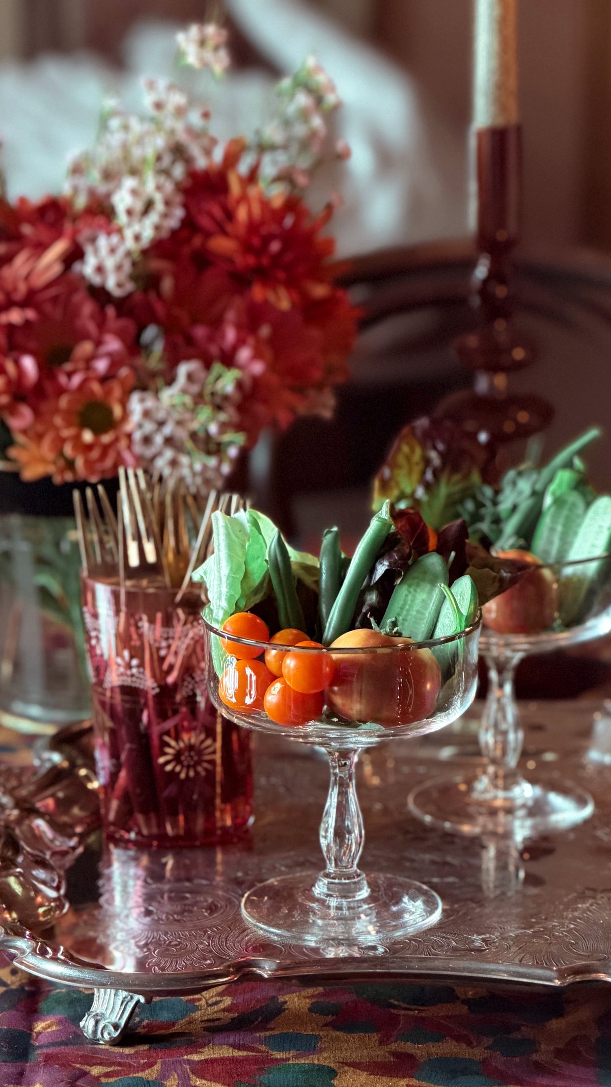 A styled tabletop scene with clear glass pedestal bowls filled with green leafy vegetables and small orange fruits, alongside a pink glass container with toothpick handles and a blurred floral arrangement in the background.