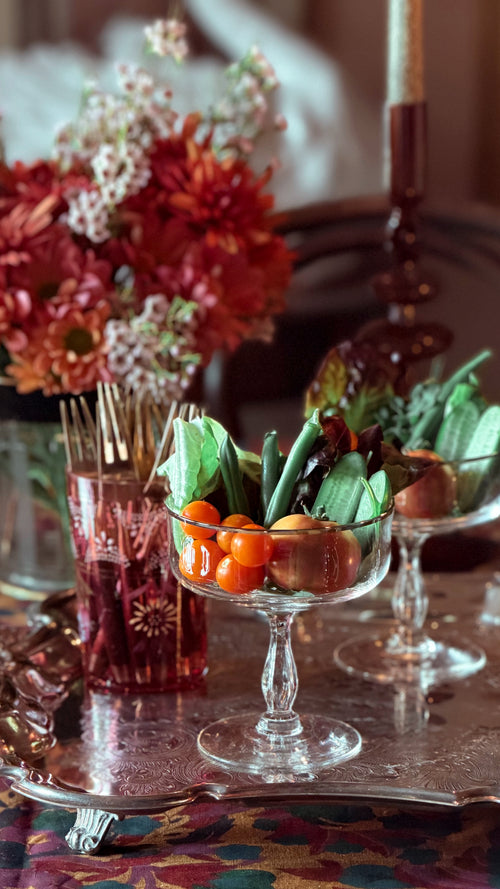 A styled tabletop scene with clear glass pedestal bowls filled with green leafy vegetables and small orange fruits, alongside a pink glass container with toothpick handles and a blurred floral arrangement in the background.