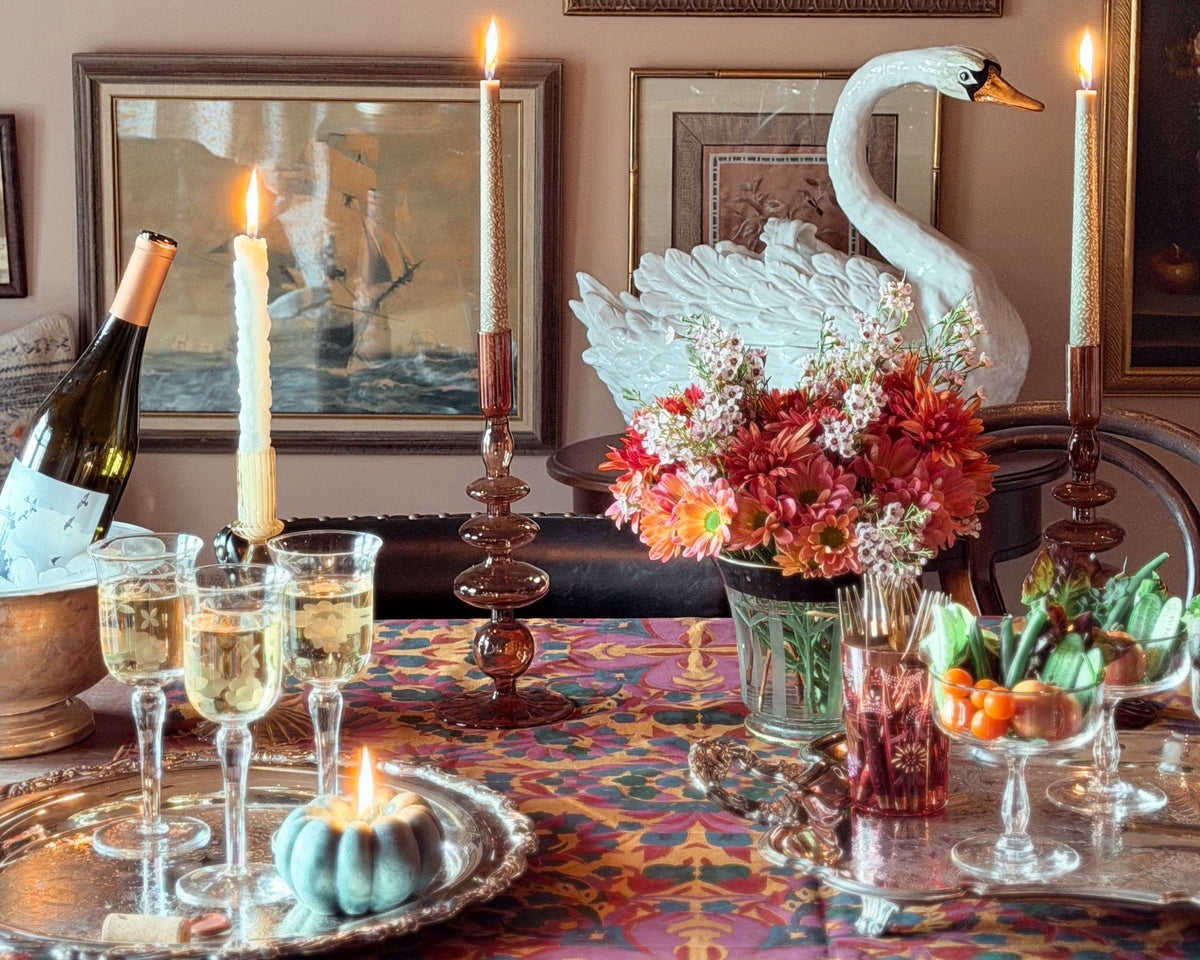 A styled dining table in a richly decorated room, featuring a colorful patterned tablecloth, lit taper candles, glasses of champagne, a floral centerpiece, and a white swan figurine against a wall covered in framed art.
