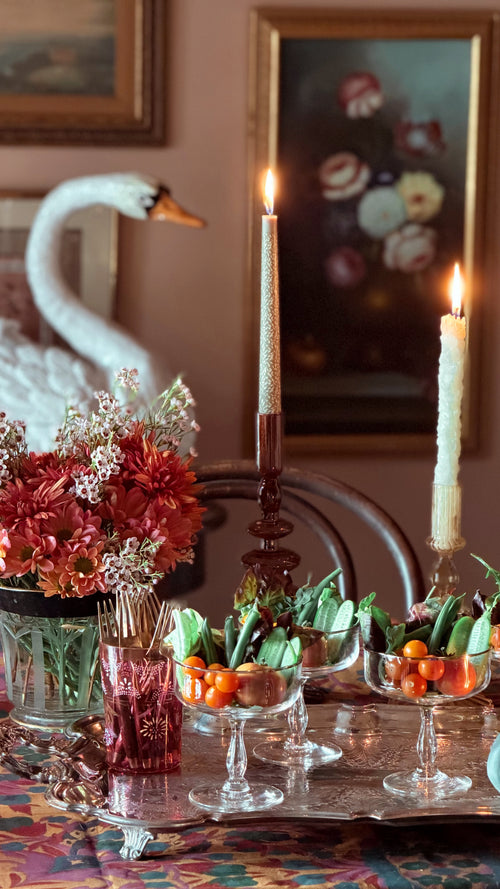 A styled dining table scene with a silver tray holding glass goblets filled with small orange fruits and green leaves, two lit taper candles in dark holders, a vase of red and white flowers, and a white swan figurine against a warm brown wall with framed artwork.