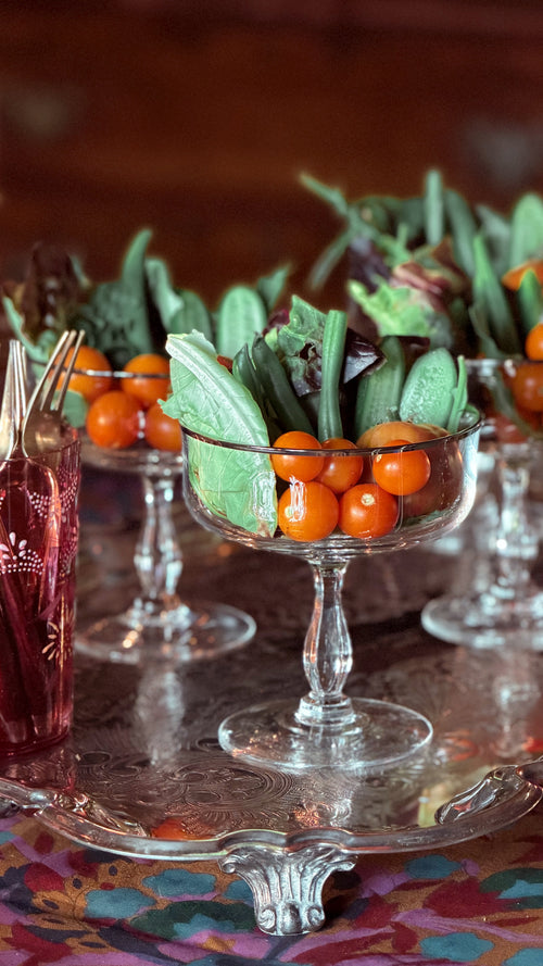 A styled tabletop arrangement with clear glass pedestal bowls containing green salad leaves and small orange berries, set on a reflective silver tray with a patterned tablecloth and a dark warm background.