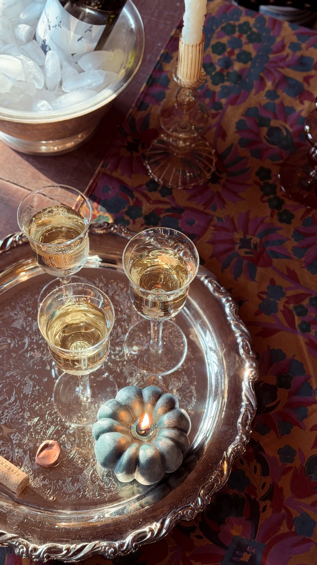 A styled tabletop scene with three small glasses of white wine on an ornate silver tray, alongside a lit candle, a cork, and a bowl of ice. The tray rests on a richly patterned purple and gold tablecloth.