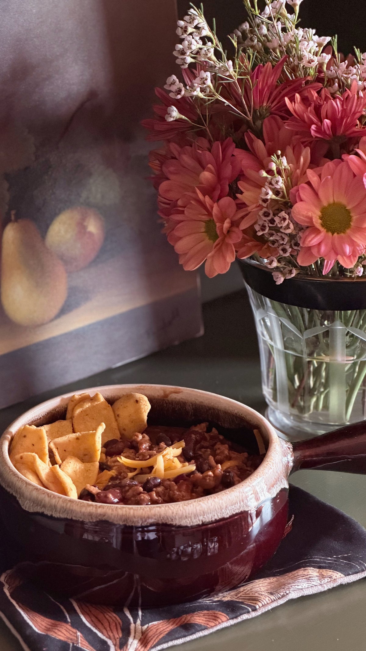 A stoneware bowl filled with chili and tortilla chips sits on a patterned cloth on a tabletop, with a vase of pink and peach flowers in the background.