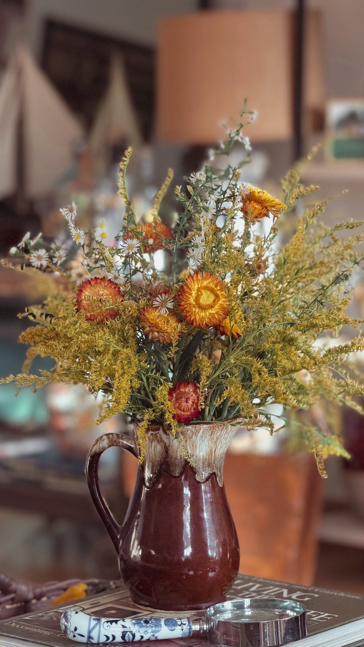 A rustic brown ceramic pitcher vase filled with a dried flower arrangement featuring yellow, orange, and white blossoms, set on a table in a warm, softly blurred indoor environment.