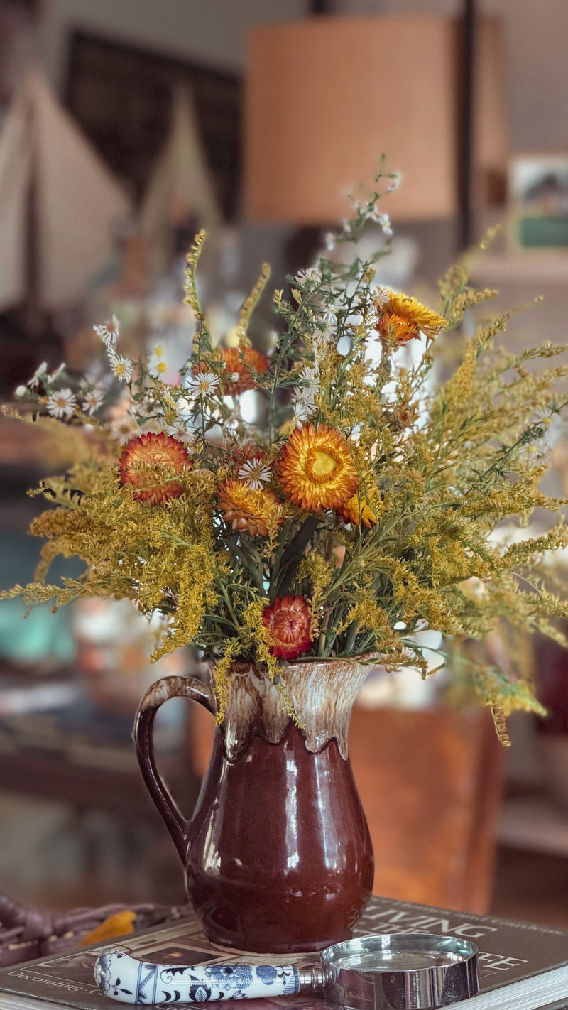 A rustic brown ceramic pitcher vase filled with a dried flower arrangement featuring yellow, orange, and white blossoms, set on a table in a warm, softly blurred indoor environment.