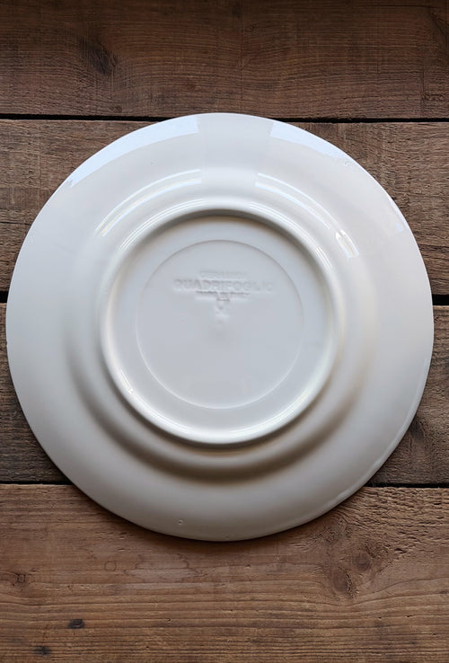 A white ceramic plate shown from the back, featuring concentric circular ridges and a faint embossed brand mark, placed on a rustic dark brown wooden surface.