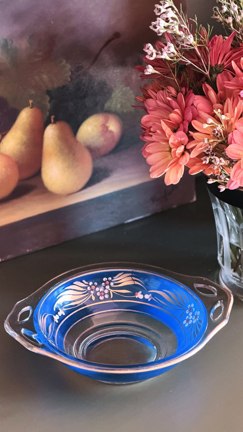 A blue glass bowl with white floral patterns and side handles, placed on a dark tabletop in a styled interior scene with flowers and fruit in the background.