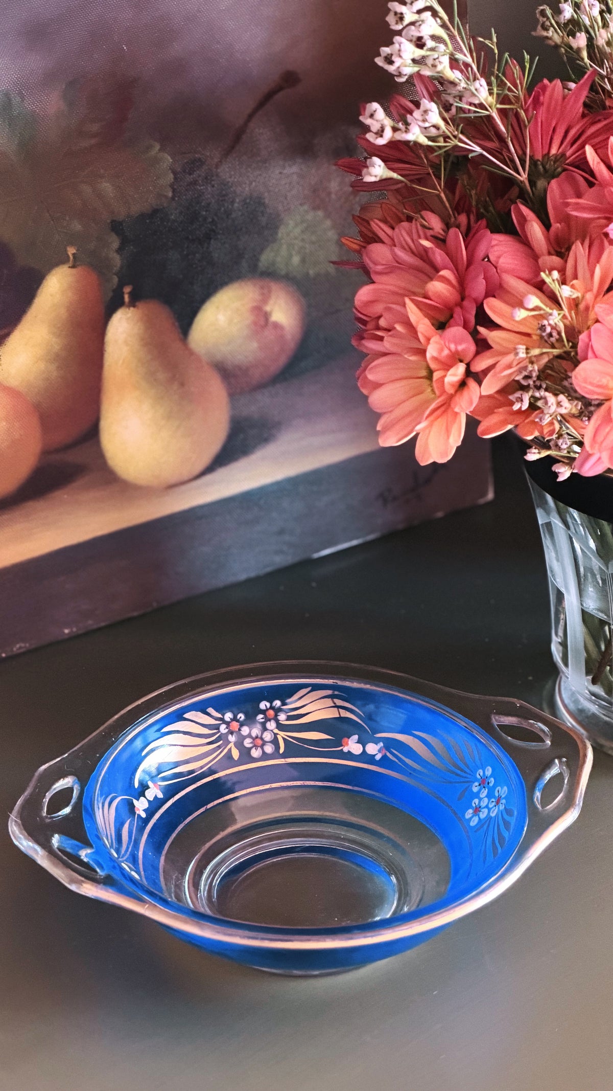 A blue glass bowl with white floral etched patterns, displayed on a dark tabletop next to a vase of pink flowers, with a still life painting of pears in the background.