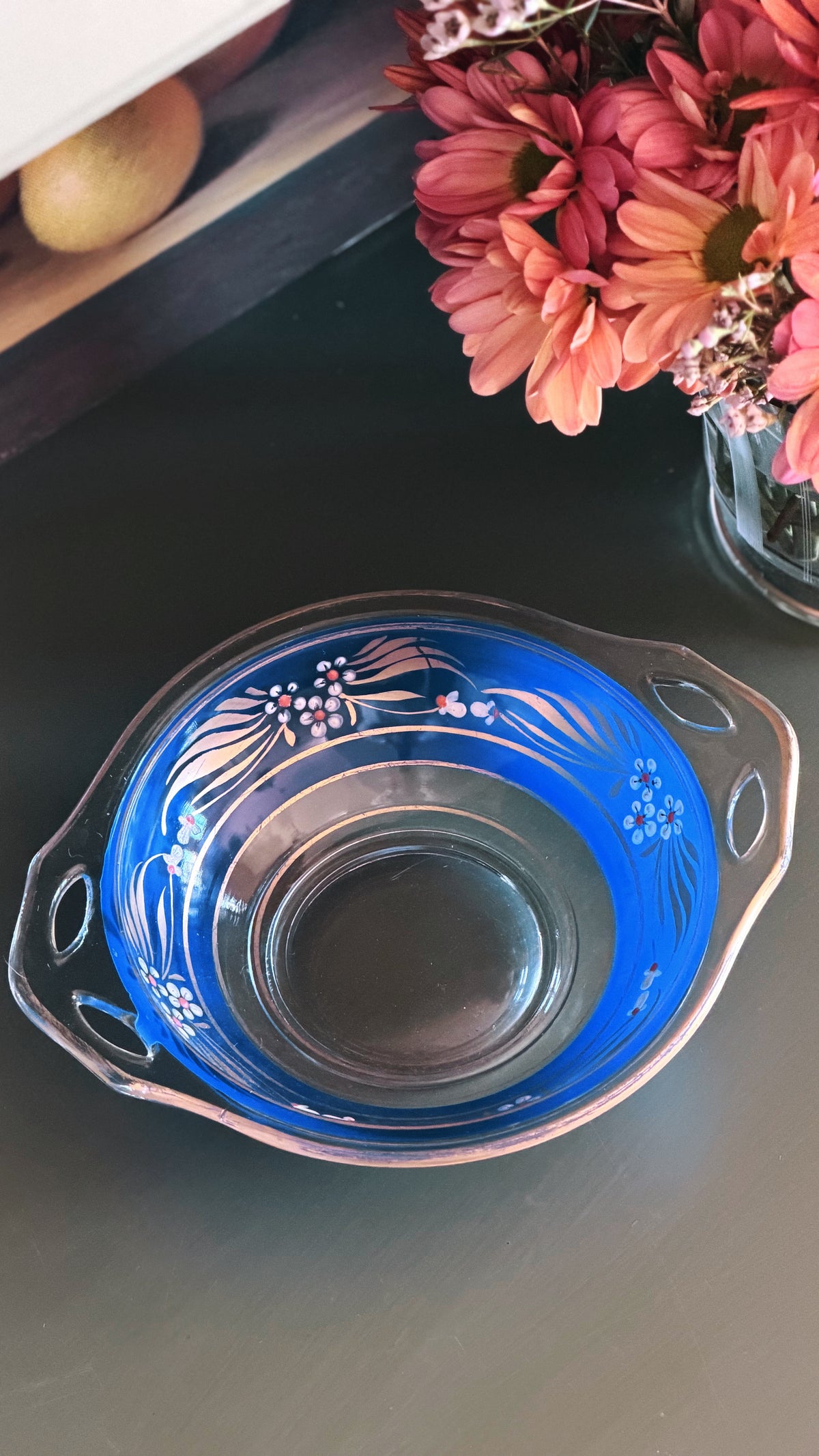 A clear glass serving bowl with a vibrant blue interior and white floral patterns, shown on a dark tabletop next to pink flowers.