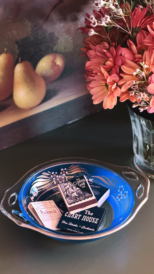 A decorative blue glass bowl with silver-toned handles sits on a dark tabletop, holding a miniature book and matchbox set. A vase of pink flowers and a framed still life with pears are visible in the background.