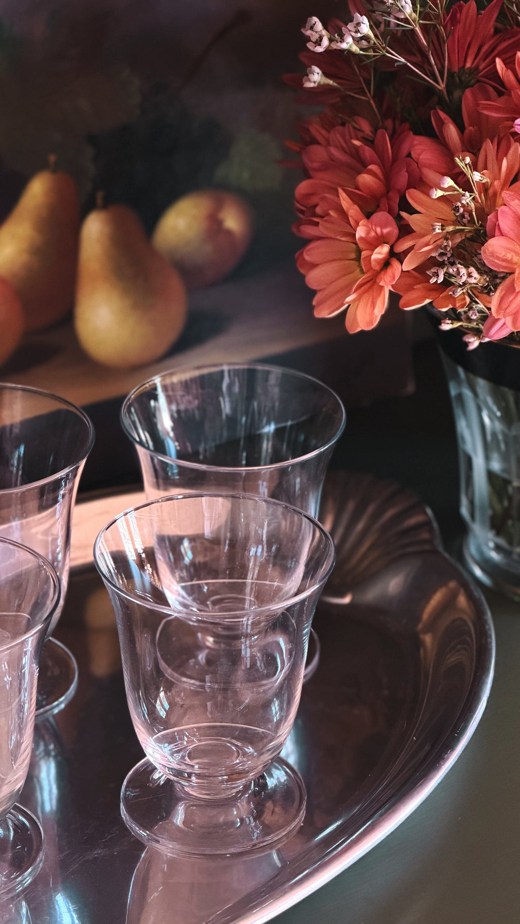 Clear glass footed cups arranged on a reflective silver tray, with a blurred floral arrangement and pears in the background.