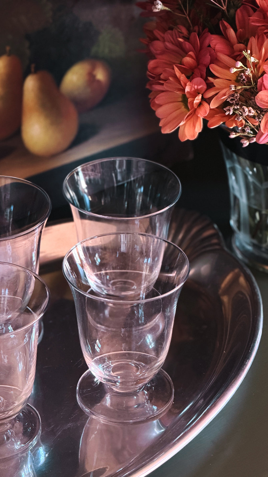 Clear glass footed cups arranged on a reflective silver tray, with warm-toned flowers and pears blurred in the background.