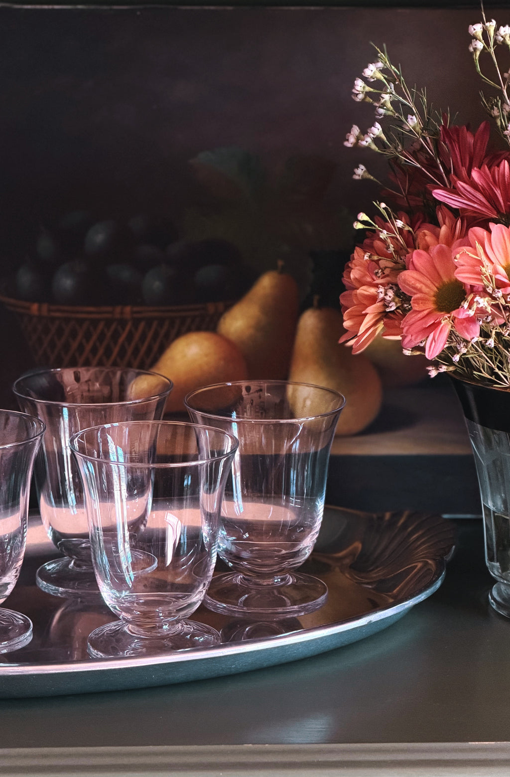 A set of clear glass stemware glasses arranged on a silver tray, with a dark background and decorative flowers and fruit in the scene.