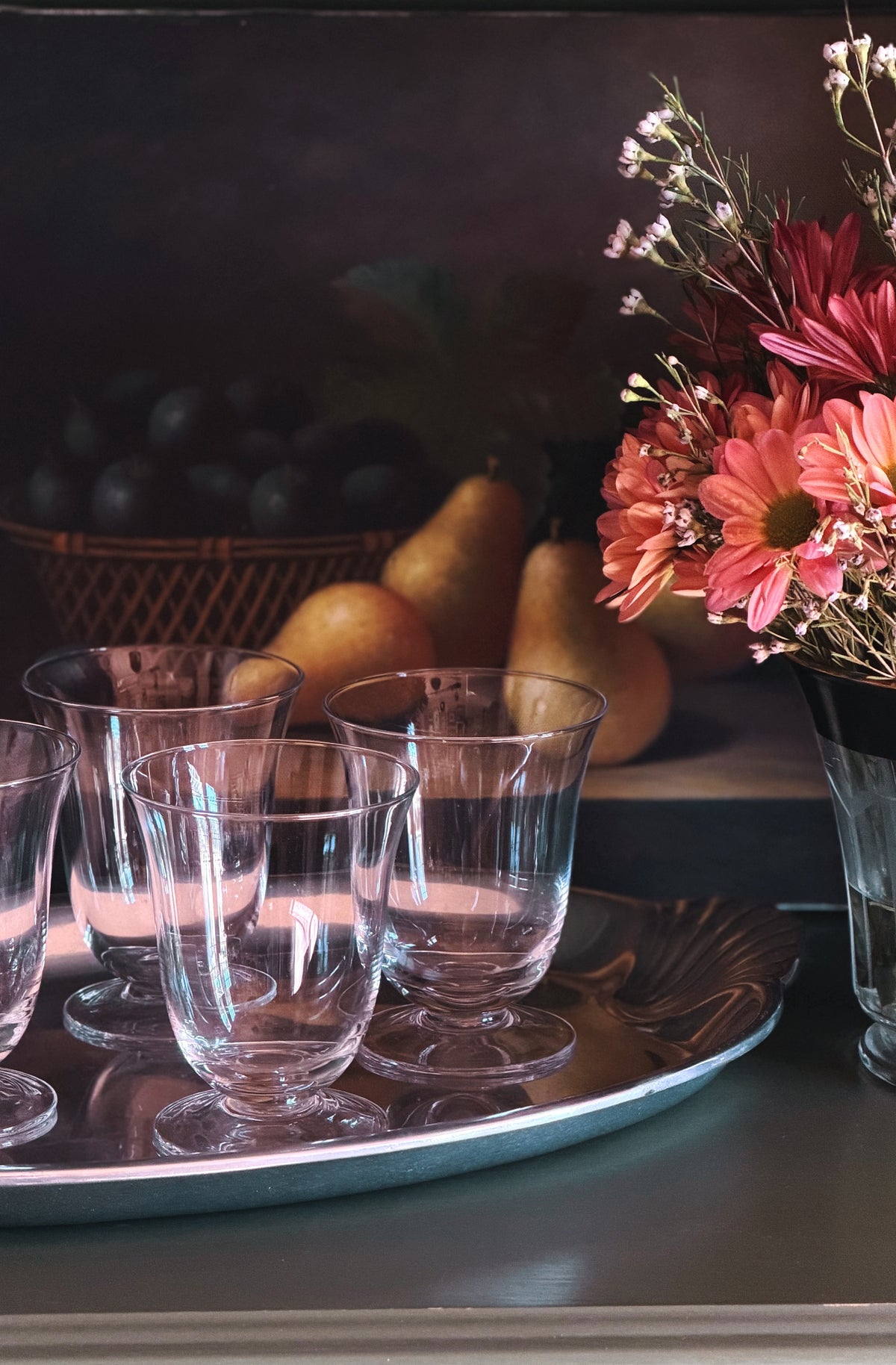 A set of clear glass stemware glasses arranged on a silver tray, with a dark background and decorative flowers and fruit in the scene.