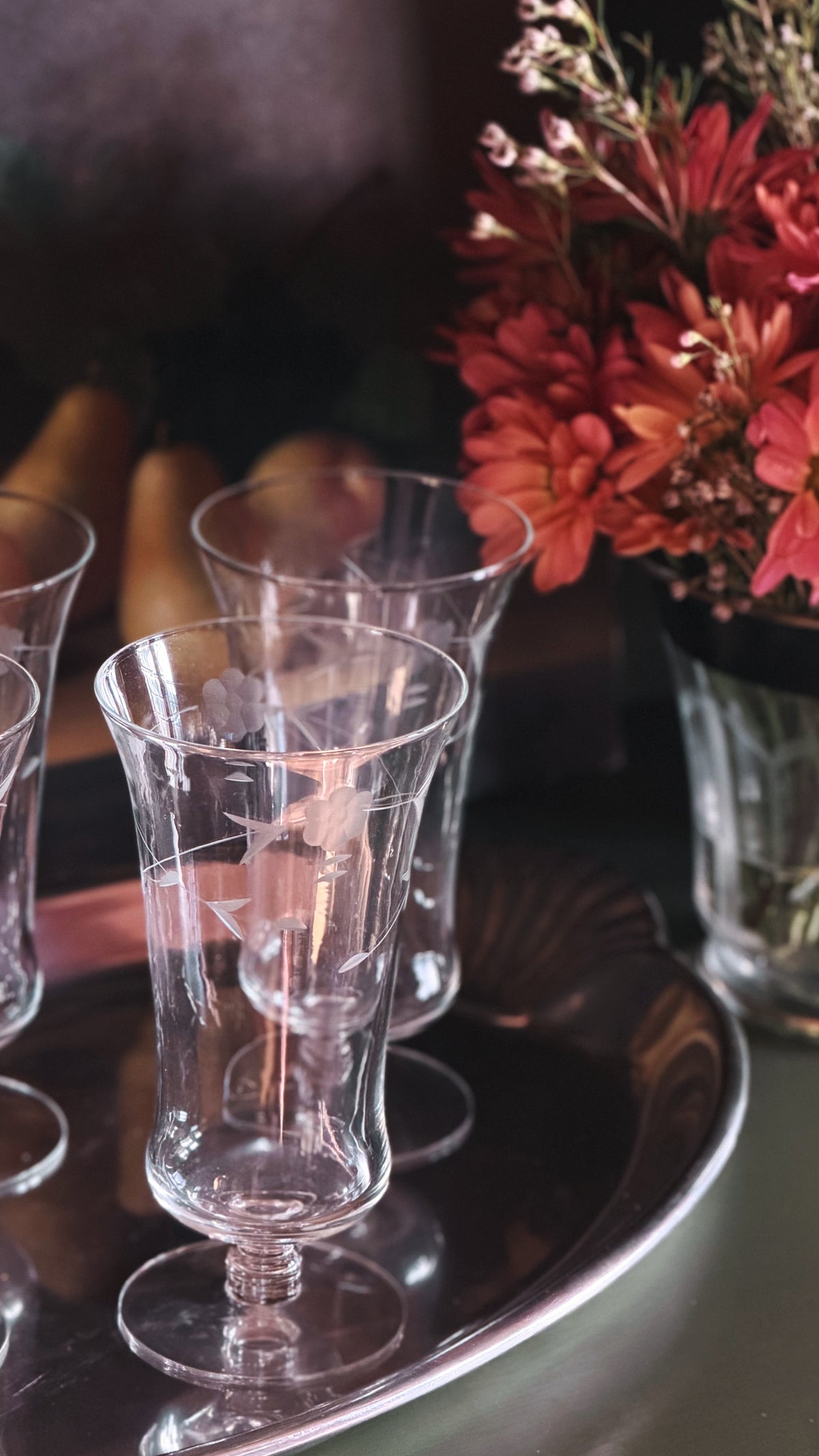 Clear glass stemware glasses arranged on a reflective dark tray, with a blurred floral arrangement in the background.