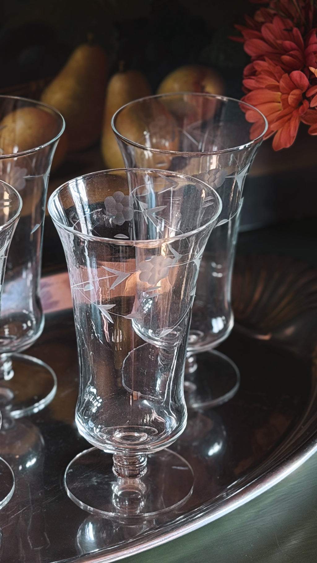 A set of clear glass tulip-shaped stemware glasses arranged on a reflective silver tray, with softly blurred pears and a red flower in the background.