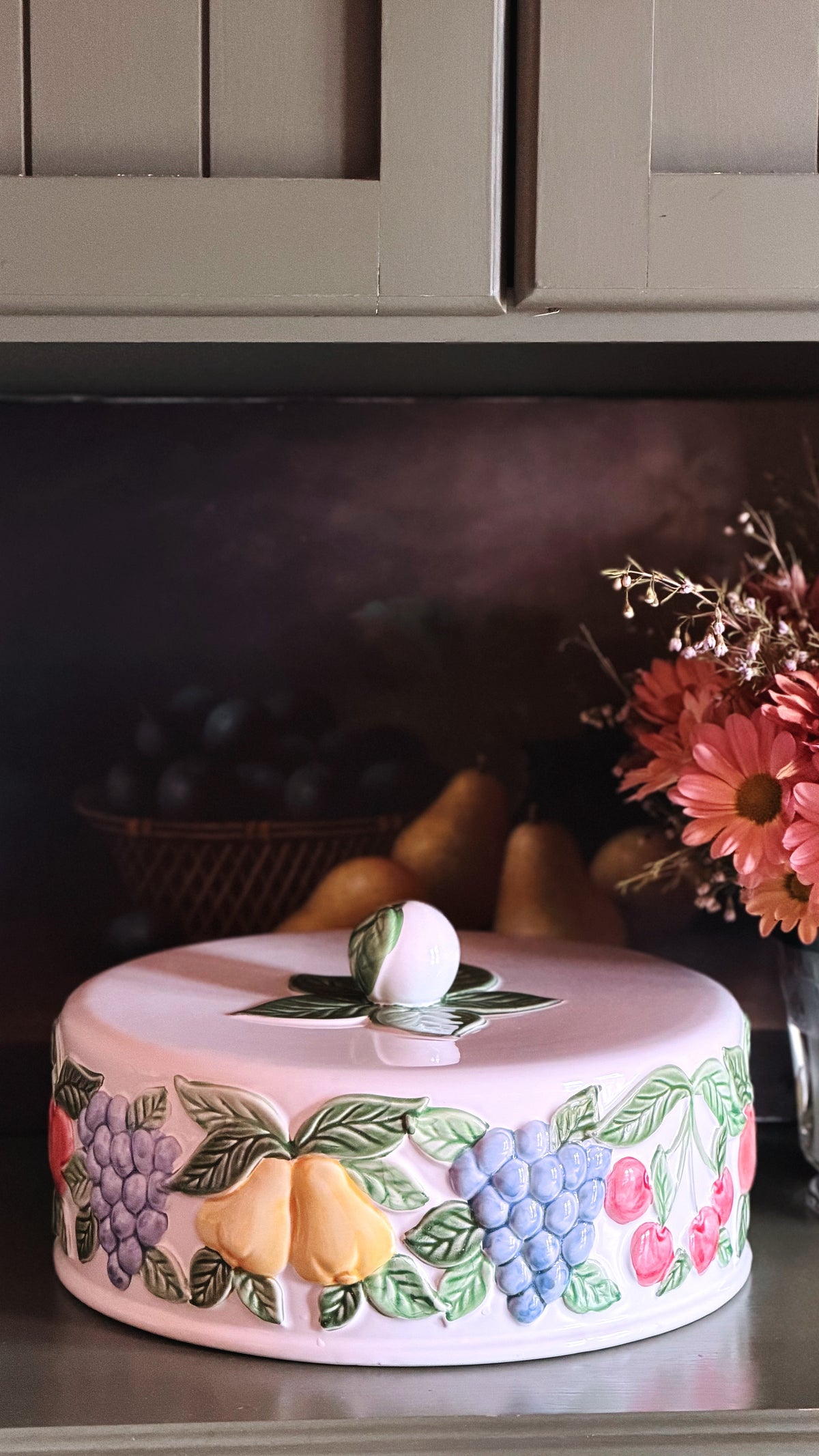 A round cake or decorative cake with a lid, decorated with 3D fruit designs including blue grapes, yellow pears, and red cherries with green leaves, displayed on a kitchen shelf.