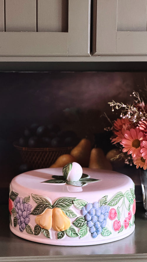 A round cake or decorative cake with a lid, decorated with 3D fruit designs including blue grapes, yellow pears, and red cherries with green leaves, displayed on a kitchen shelf.