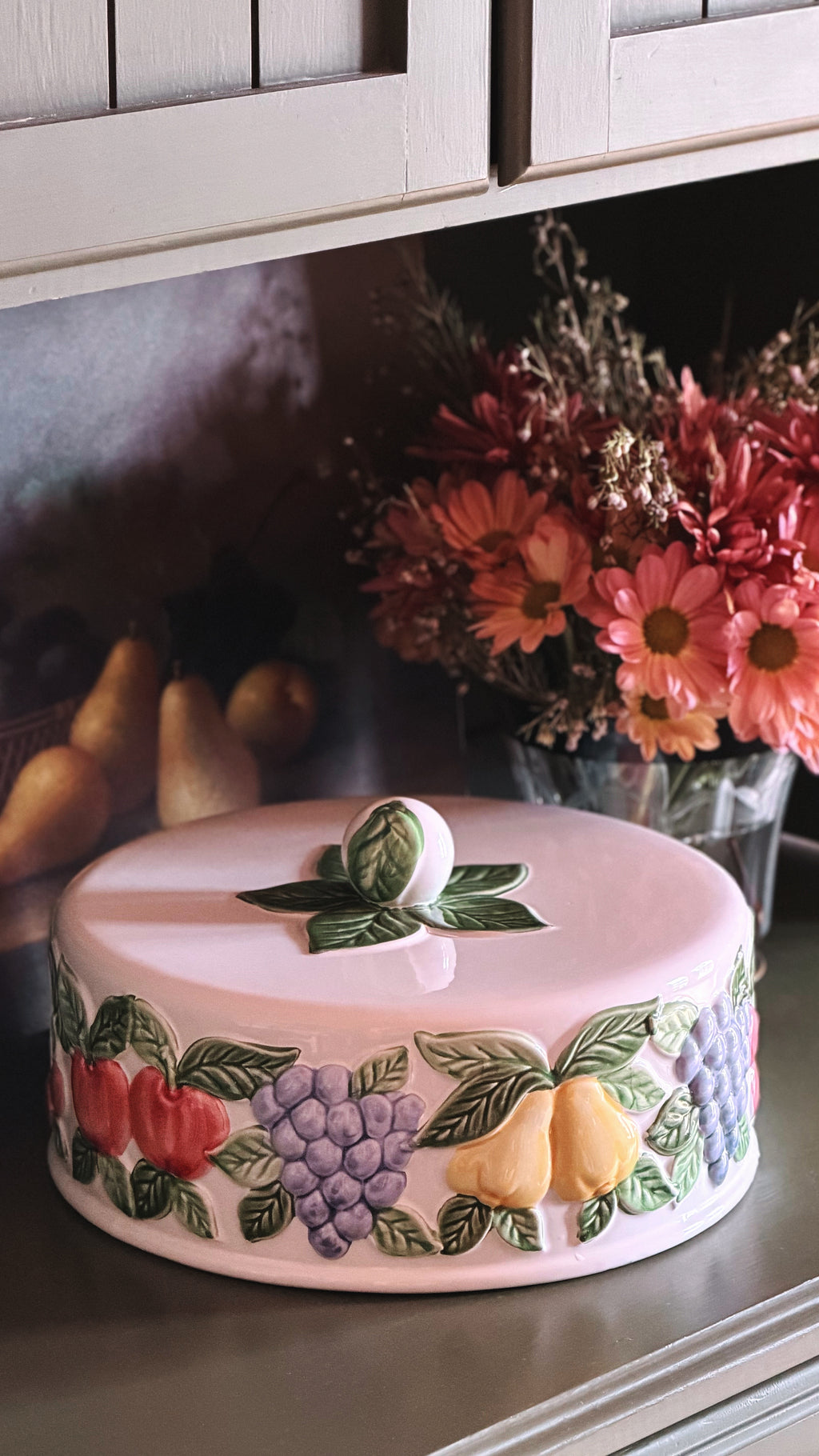 A round ceramic cake lid, decorated with colorful raised fruit motifs including red apples, purple grapes, and yellow pears, shown on a shelf with flowers in the background.